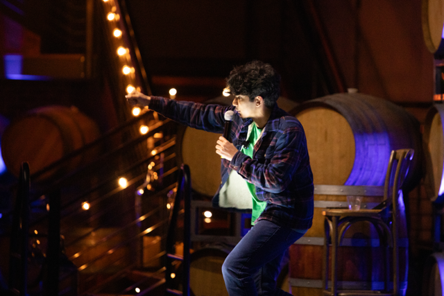 Comedian performing stand-up on stage with a microphone, set against a backdrop of wooden barrels and ambient lighting.