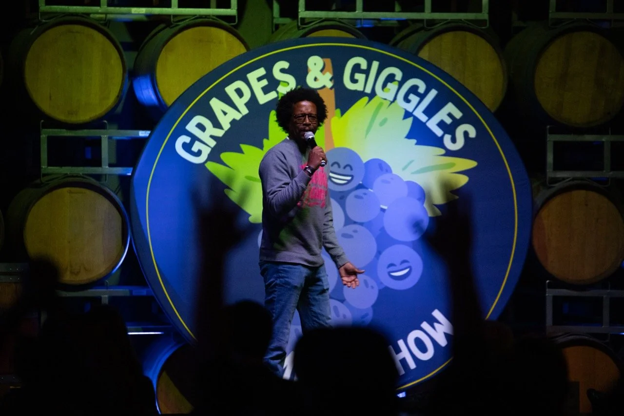 A comedian performing on stage with a "Grapes & Giggles" backdrop, surrounded by wine barrels.