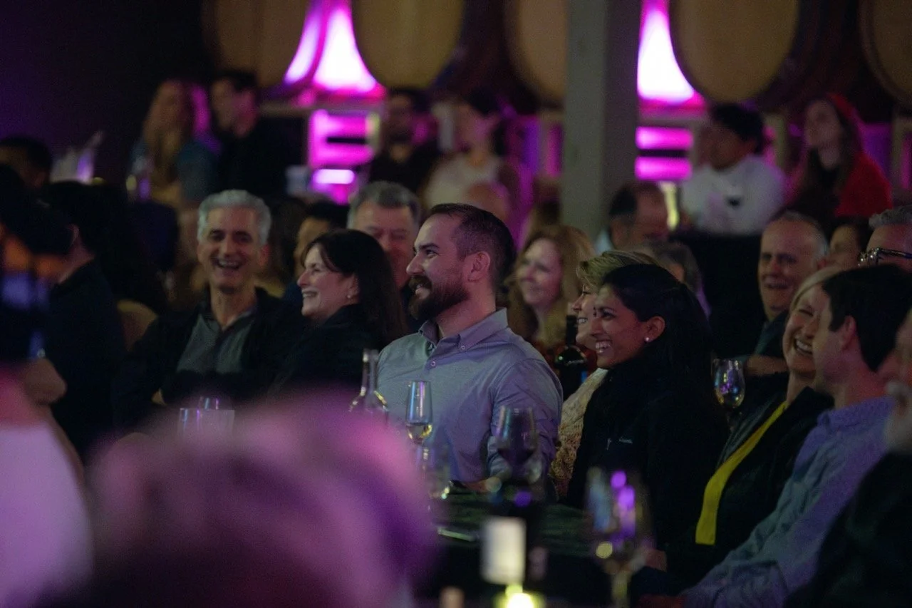 Group of people laughing at an event, with wine glasses on the table, and purple lighting in the background.