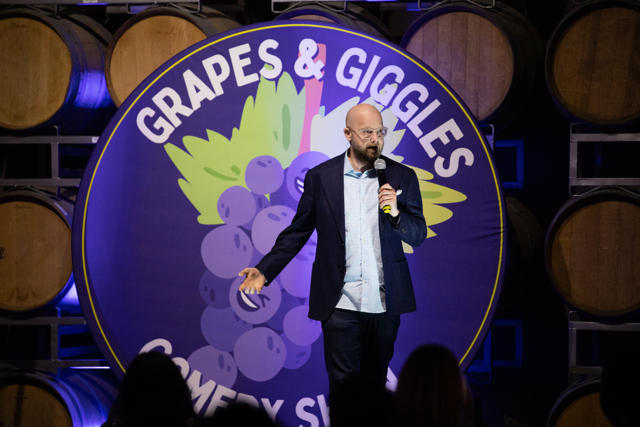 Comedian performing at "Grapes & Giggles Comedy Show" in front of a logo with a grape bunch design, surrounded by wine barrels.