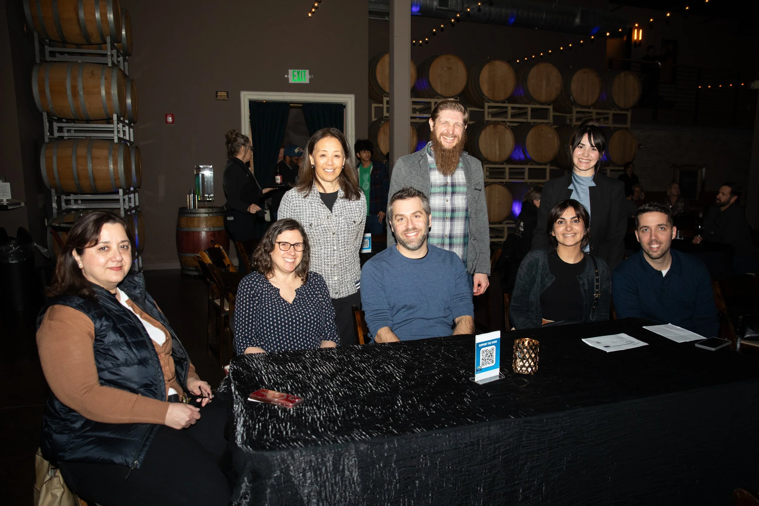 Group of people smiling around a table at an indoor event, with wine barrels in the background.