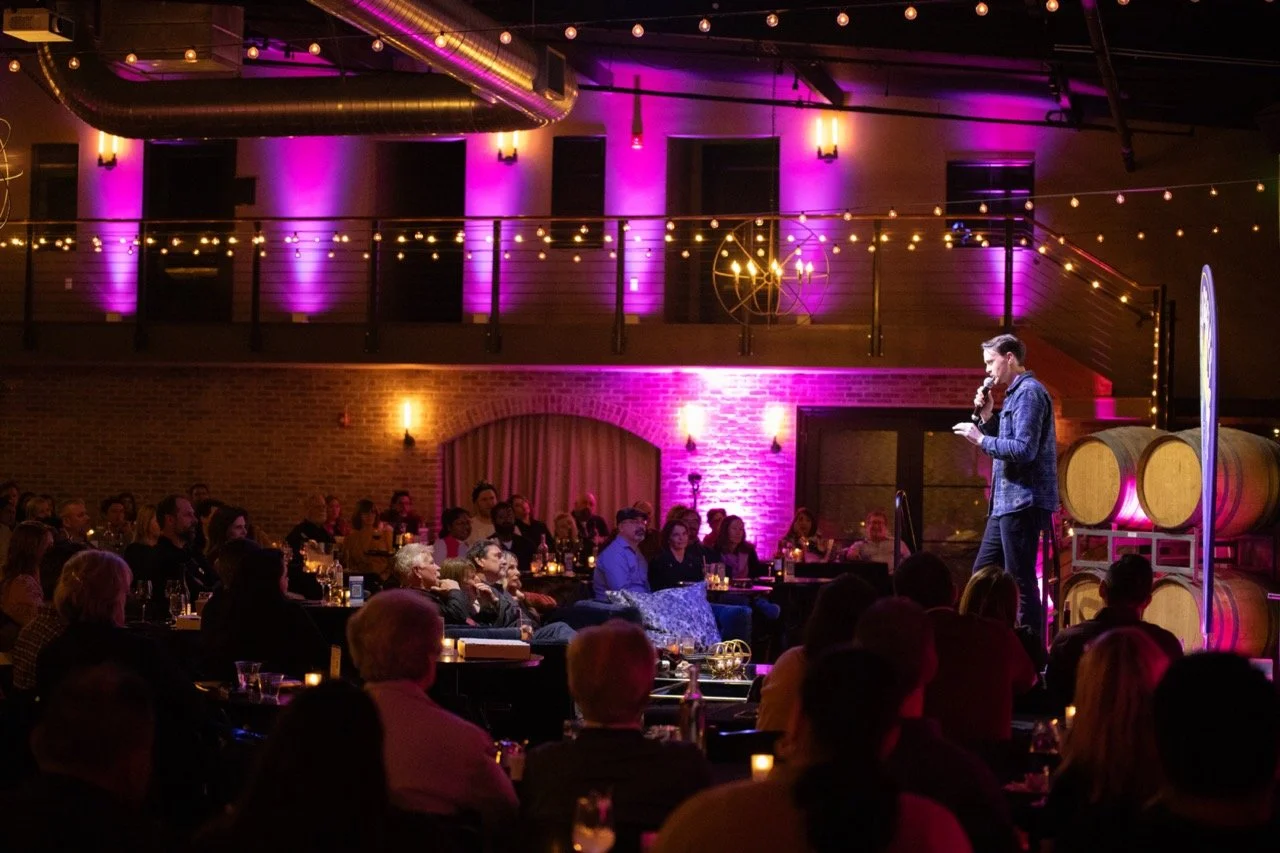 A stand-up comedian performing on stage in a dimly lit venue with purple lighting, an audience seated at tables watching, and a backdrop of wine barrels and exposed brick walls.
