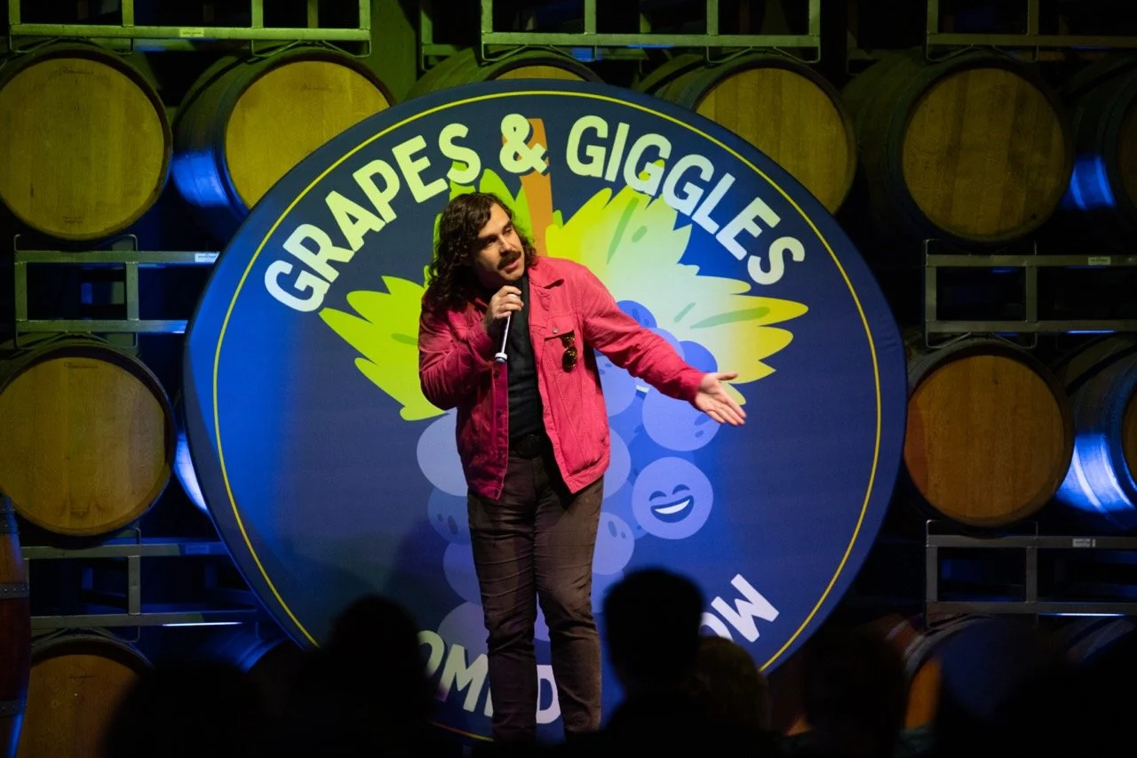 Comedian performing at "Grapes & Giggles" comedy show with wooden barrels in the background.