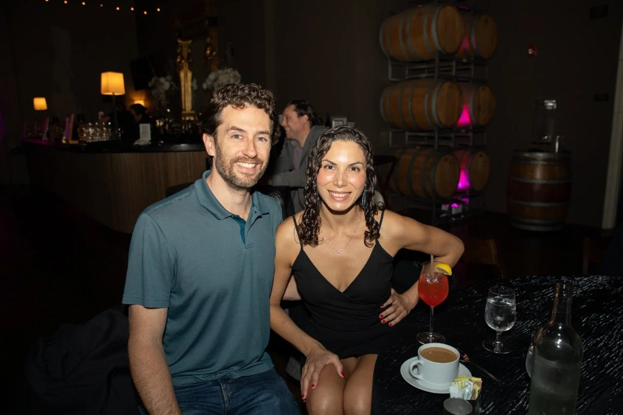 A smiling couple seated at a table in a dimly lit bar with drinks. The woman is wearing a black dress, and the man is wearing a blue polo shirt. Behind them, there is a bar counter and stacks of wooden barrels with purple lighting.