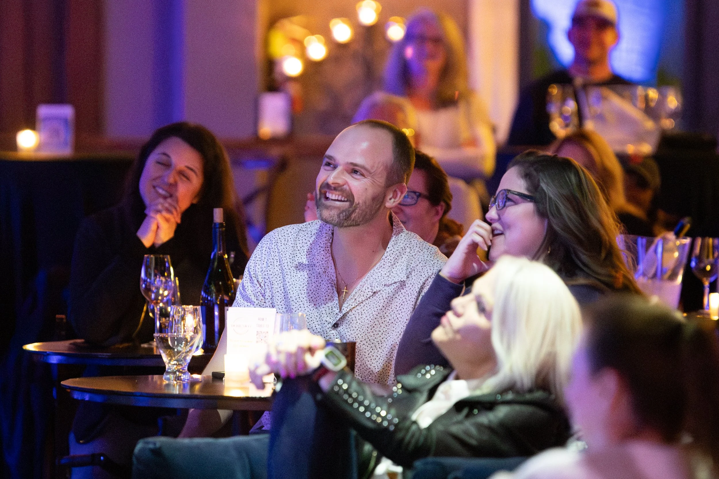A group of people sitting at tables, smiling and enjoying a performance or event in a dimly-lit venue. Wine glasses and bottles are visible on the tables.