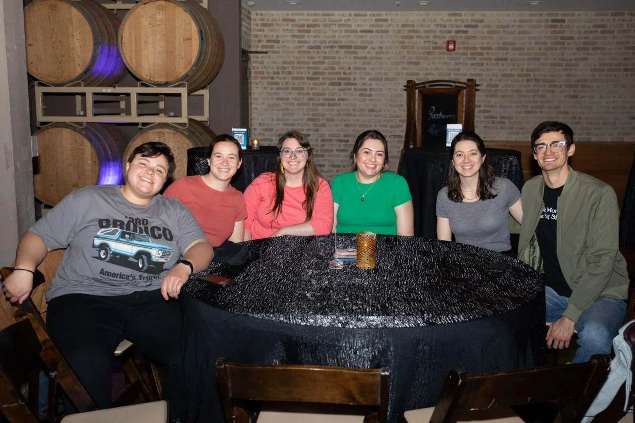 Group of six people sitting at a round table with black tablecloth, smiling; wine barrels in the background.