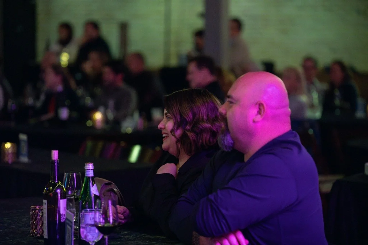 People sitting at a dimly lit event, smiling and watching a performance, with drinks on the table.