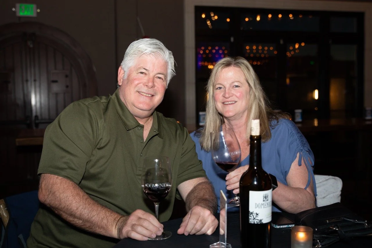 A man and woman seated at a table, smiling, holding wine glasses, with a bottle of wine on the table. The setting appears to be a restaurant or wine tasting venue with dim lighting in the background.