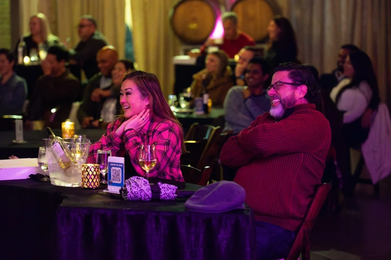 People sitting at a table in a comedy or entertainment venue, smiling and laughing, with drinks and candles on the table.