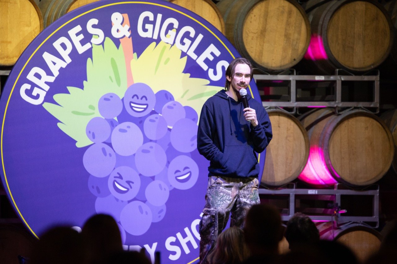 Comedian performing at a comedy show with a "Grapes & Giggles" logo and wine barrels in the background.