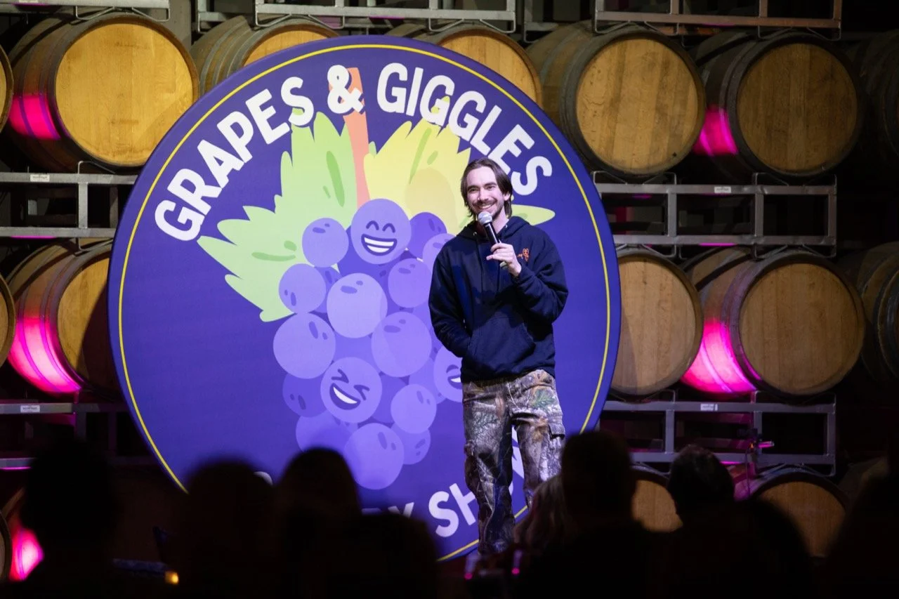 Comedian performing in front of "Grapes & Giggles" sign at a winery.