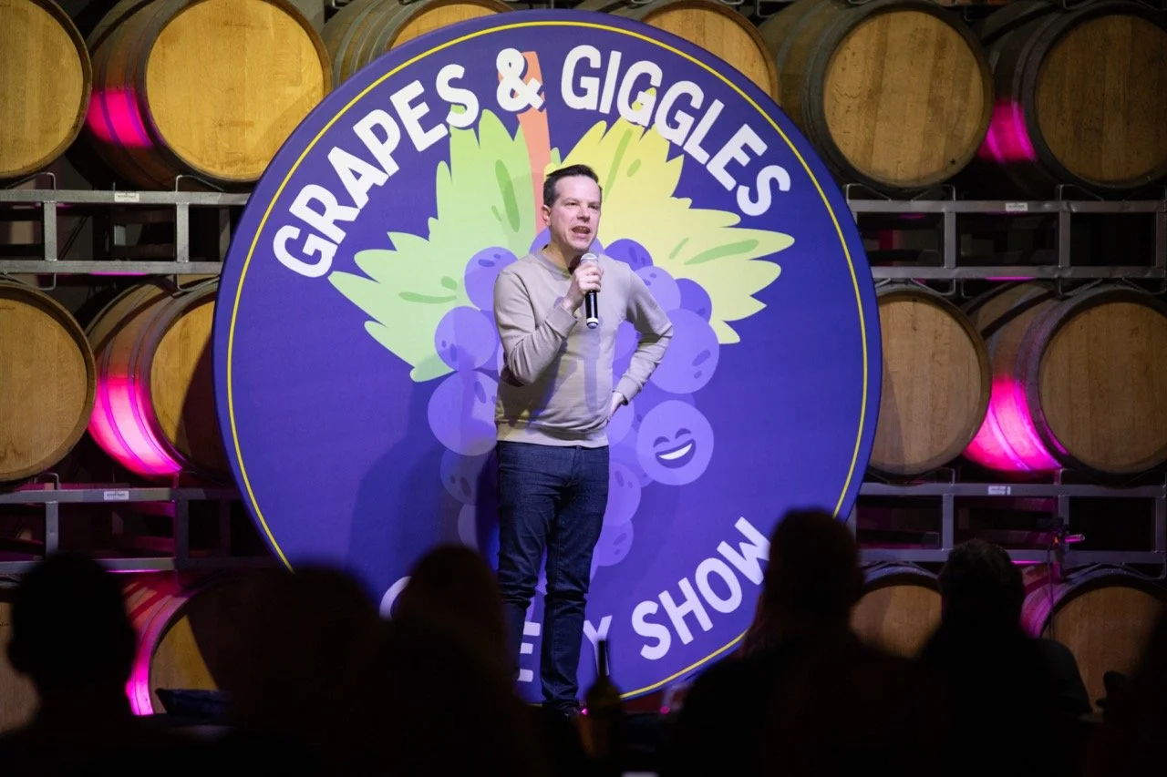 Comedian performing at "Grapes & Giggles" comedy show with wine barrels in the background.