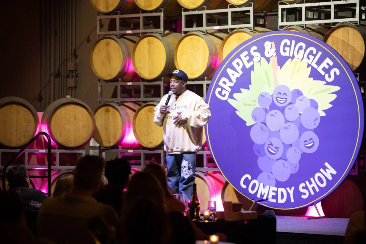 Comedian performing at Grapes & Giggles Comedy Show in a winery with wine barrels in the background.
