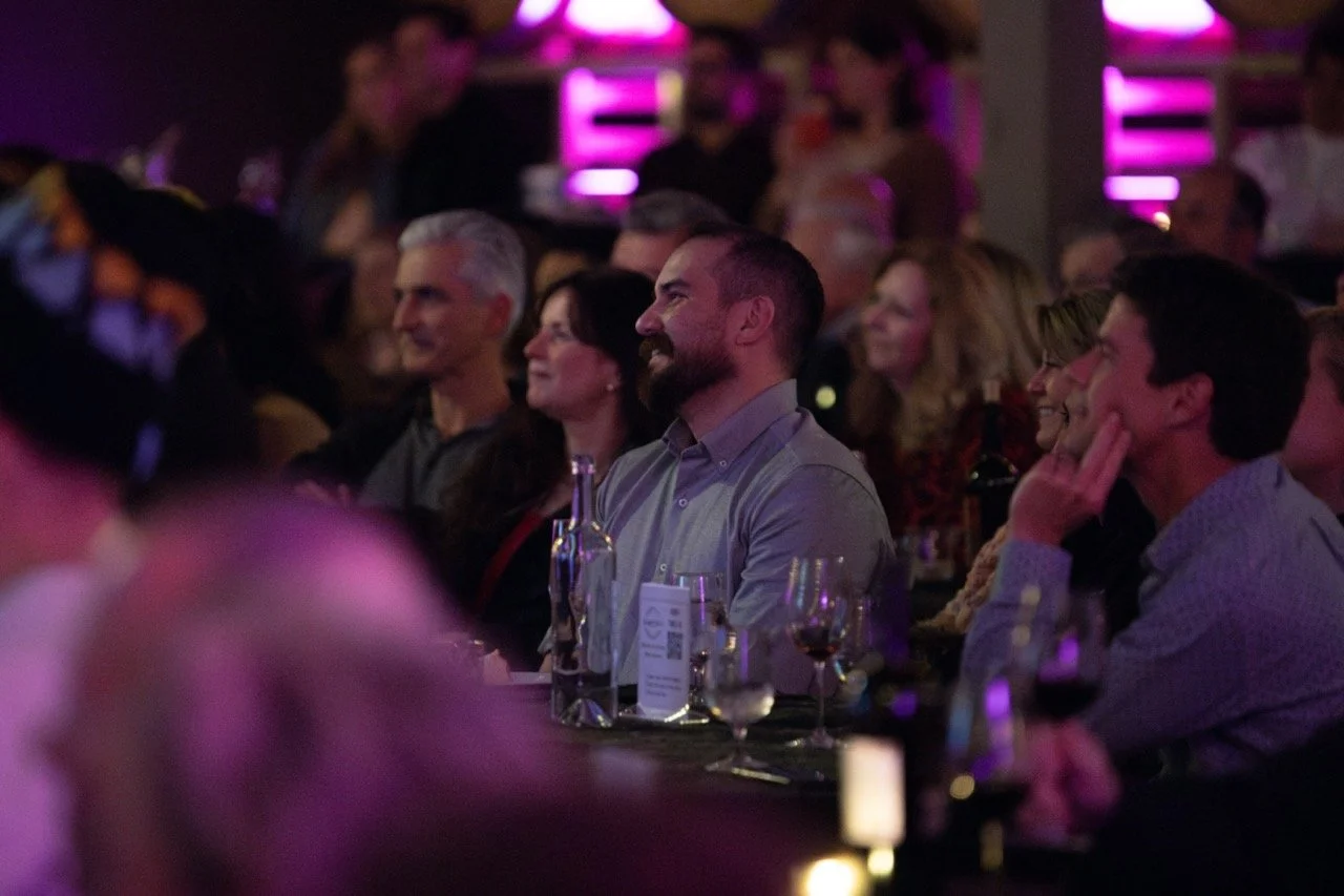 Audience seated in a dimly lit venue with pink lighting, attentively watching an event. People are smiling and engaged, with wine glasses and a menu on the table.