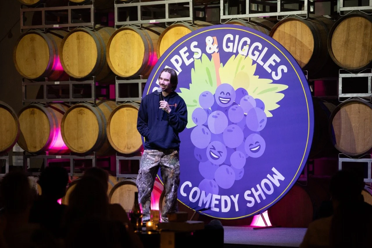 Comedian performing at Grapes & Giggles Comedy Show with wine barrels in background.