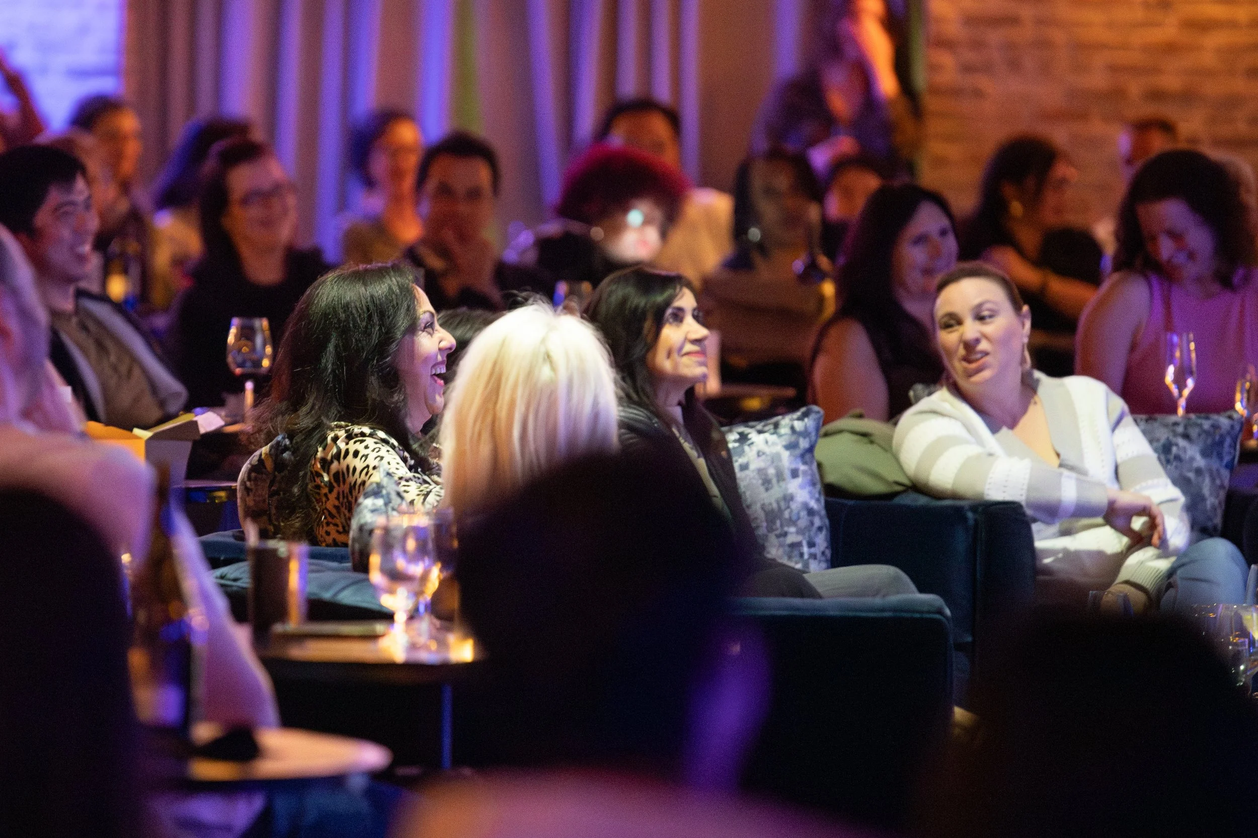 Audience enjoying a comedy show in a dimly lit club, people laughing and smiling, with drinks on the tables, seating in rows.