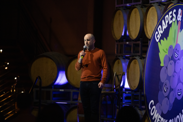 A stand-up comedian performing on stage with a microphone at a comedy show in a venue with wine barrels and a sign that reads 'Grapes & Comedy Show.'