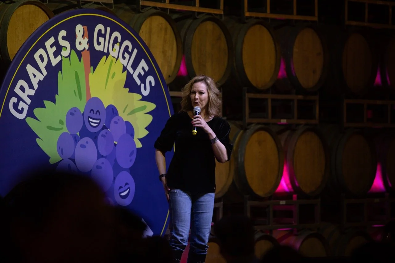 Comedian performing on stage at "Grapes & Giggles" event with wine barrels in the background.