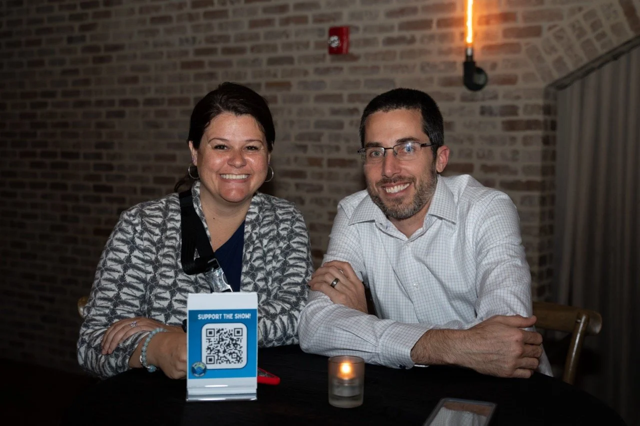 Two people smiling at a table with a "Support the Show" QR code sign
