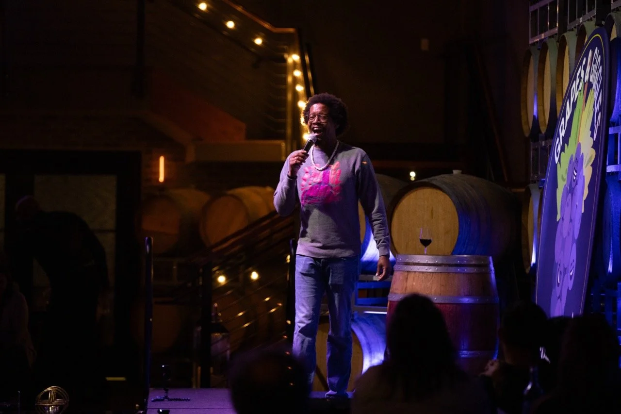 Comedian performing on stage with microphone in hand, wine barrels and audience visible, low lighting.