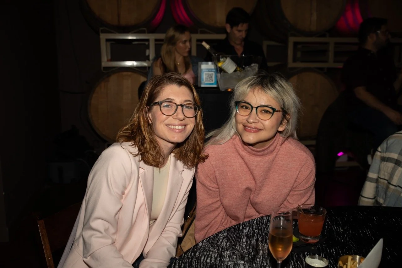 Two women smiling at a table in a dimly lit venue with barrels in the background, drinks in front of them.