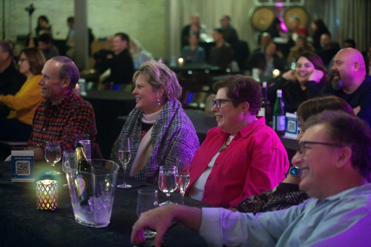 Audience enjoying a live event, seated at tables with drinks, smiling and laughing.