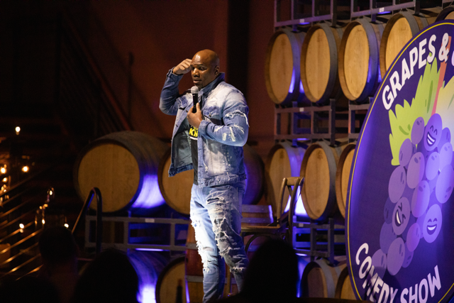 Comedian performing at Grapes & Comedy Show next to wine barrels and event sign.