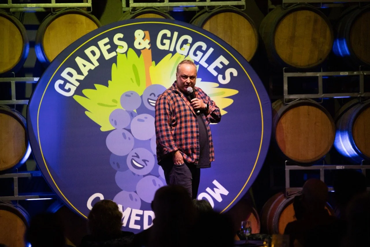 Comedian performing on stage in front of a sign that reads 'Grapes & Giggles Comedy Show,' with wine barrels in the background.