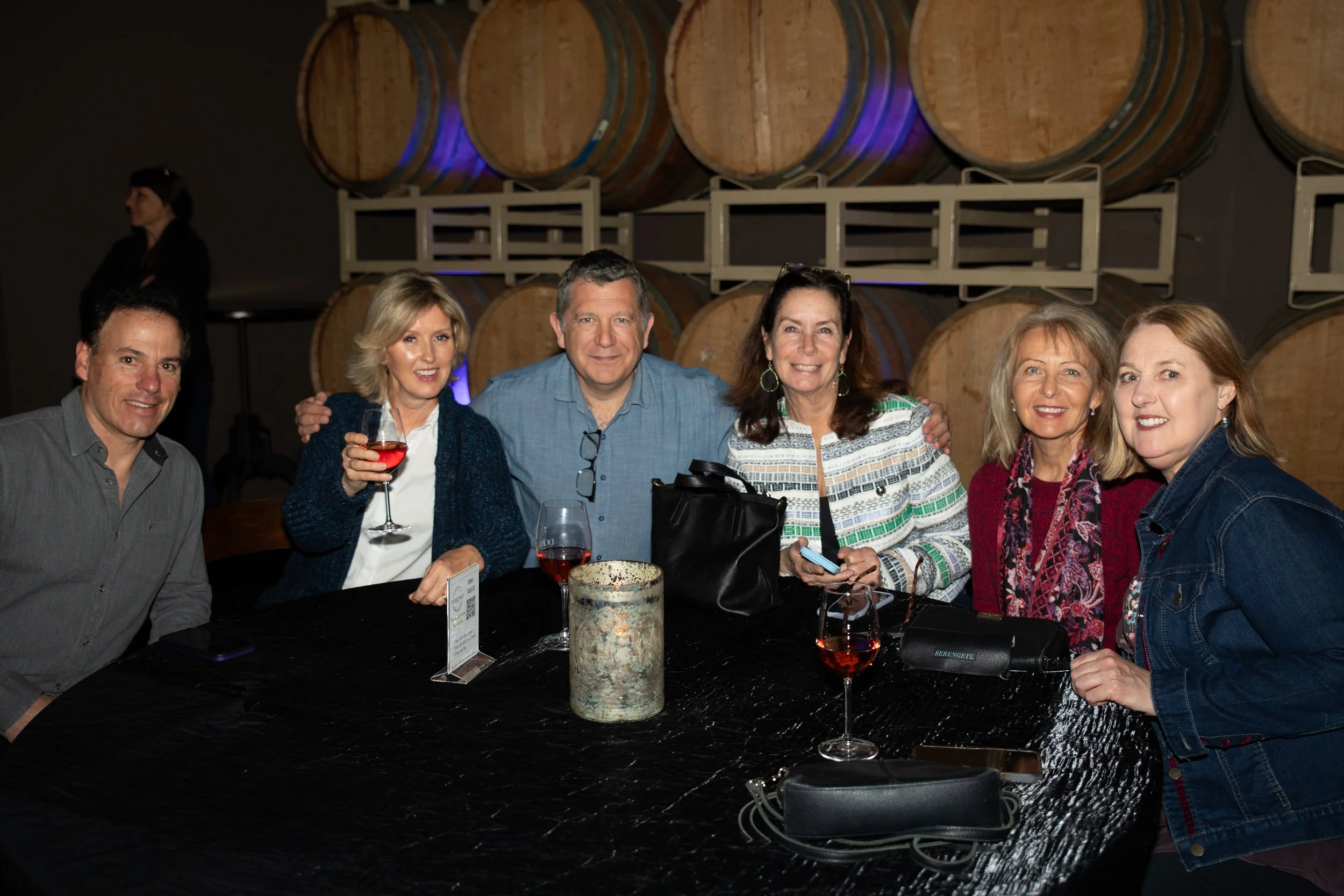 Group of six people at a table with wine glasses, in front of wine barrels, smiling at a winery.