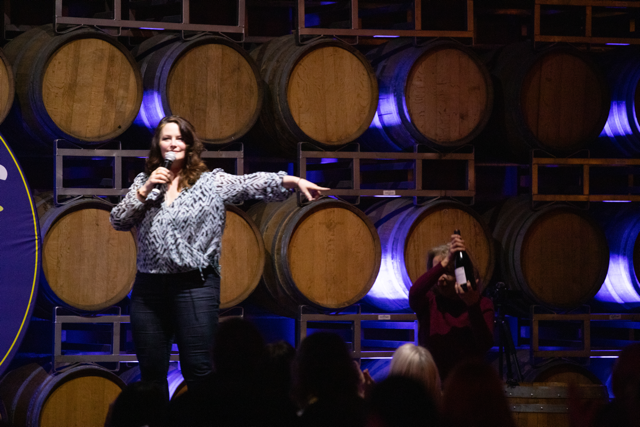 Woman speaking on stage in a winery, holding a microphone, with wine barrels in the background and an audience watching.