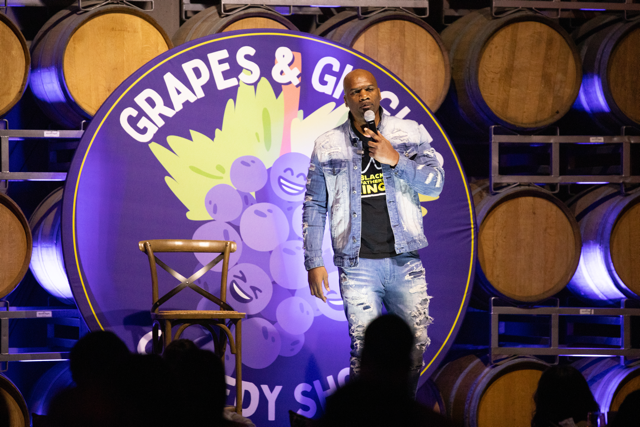 Comedian performing at "Grapes & Giggles" comedy show in front of wine barrels and a purple sign with cartoon grapes.