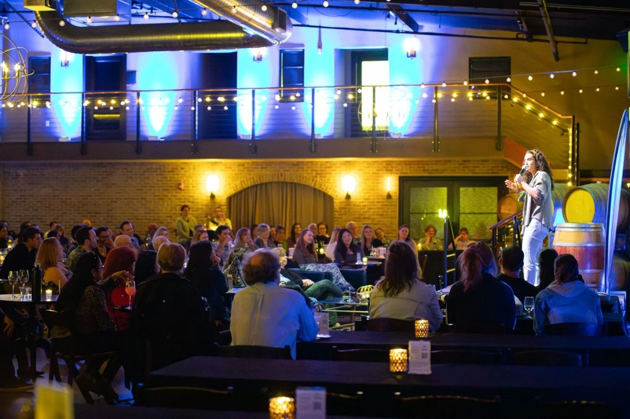 A stand-up comedian performing on stage in front of an audience at a dimly lit comedy club with blue and yellow lighting, brick walls, and wine barrels as decor.
