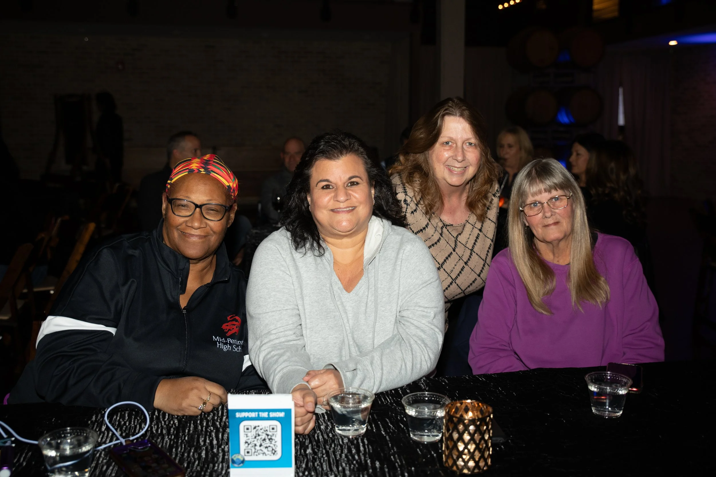 Four women sitting at a table with drinks, in a dimly lit venue, smiling for the camera.