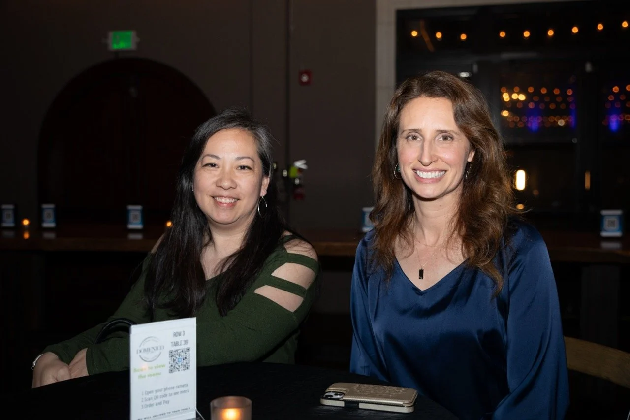 Two women sitting at a table in a dimly lit venue, smiling at the camera. A QR code menu sign and a smartphone are visible on the table.