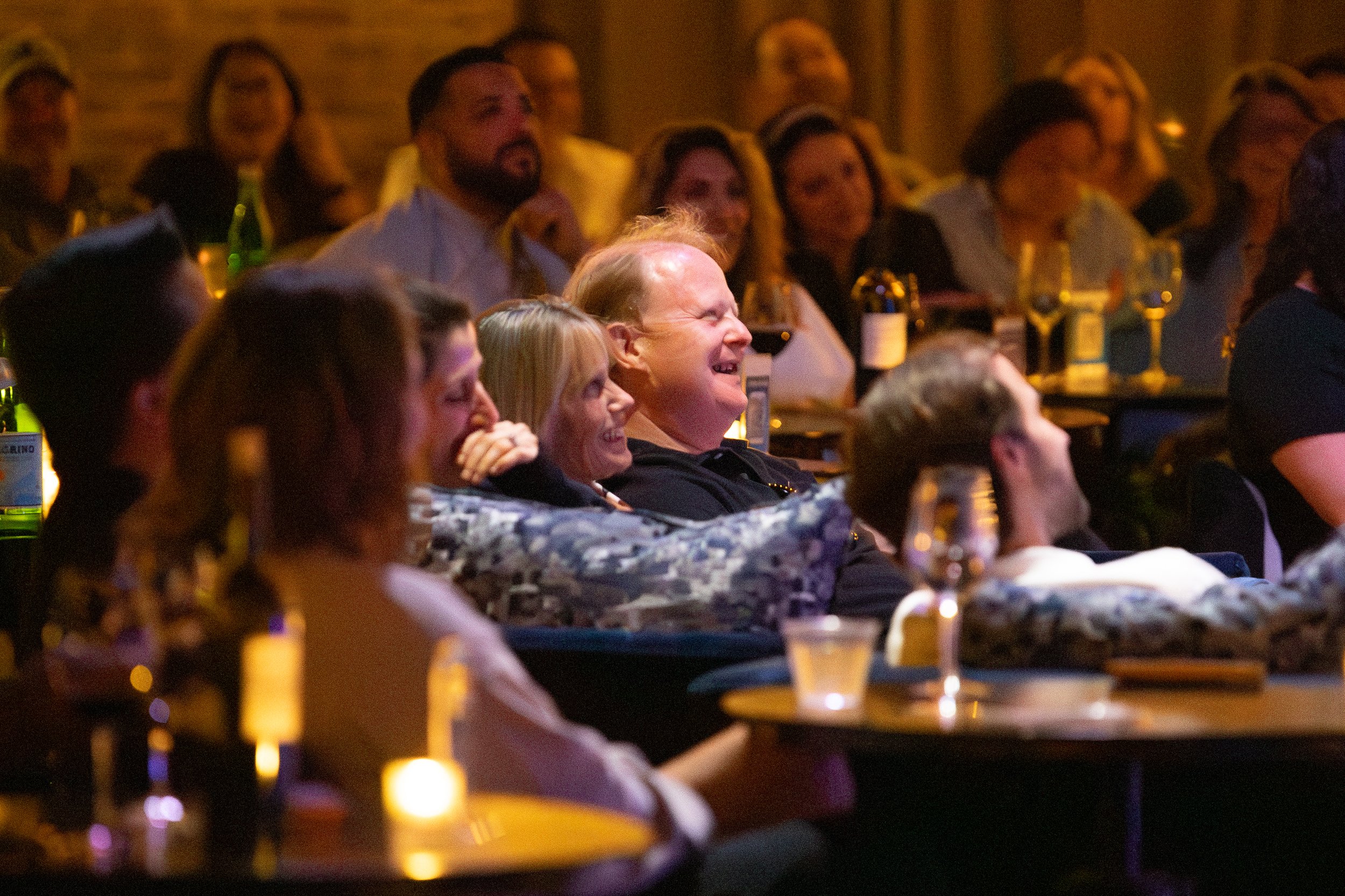 Audience laughing at a comedy show, seated at tables with drinks and candlelight in a dimly lit venue.