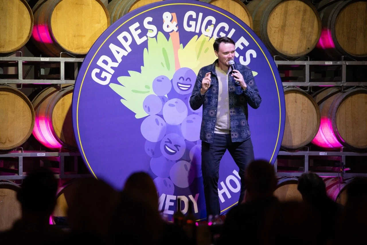 Comedian performing on stage at Grapes & Giggles comedy show with barrels in the background.