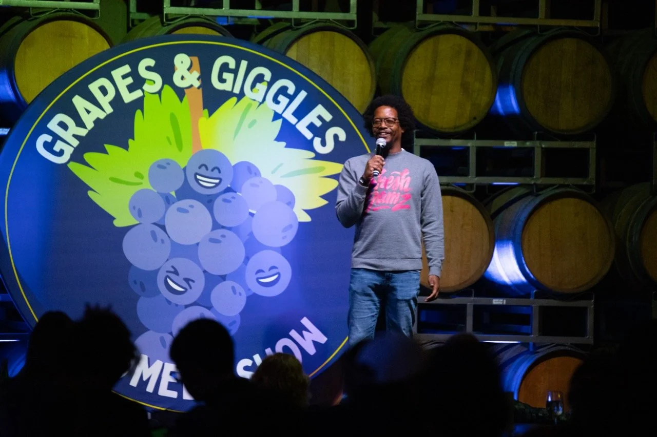 Comedian performing at 'Grapes & Giggles' comedy show, standing in front of wine barrels and a large logo with smiling grapes.