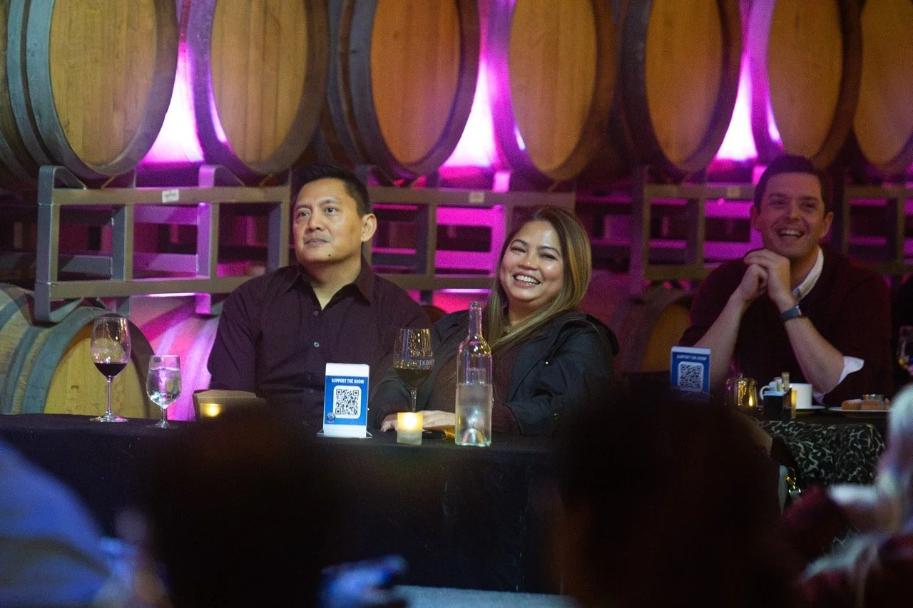 People sitting at a table with glasses of wine in a room with wooden barrels and purple lighting.