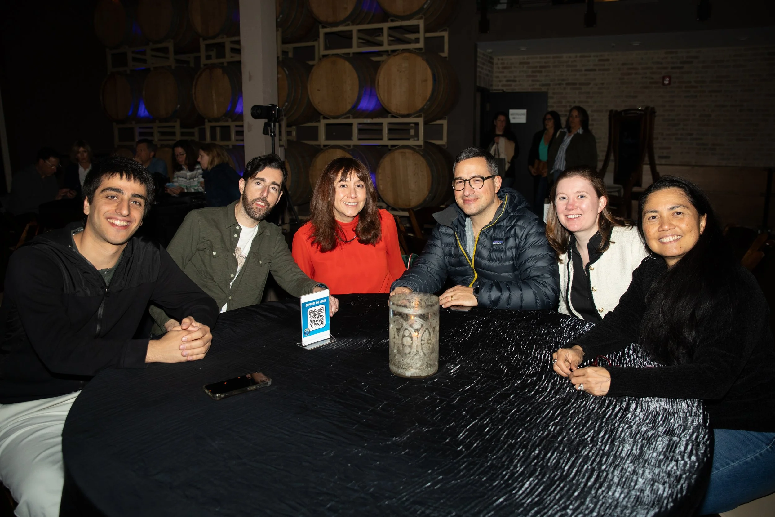 Group of six people seated at a table in a dimly lit room with barrels in the background, smiling and posing for a photo.