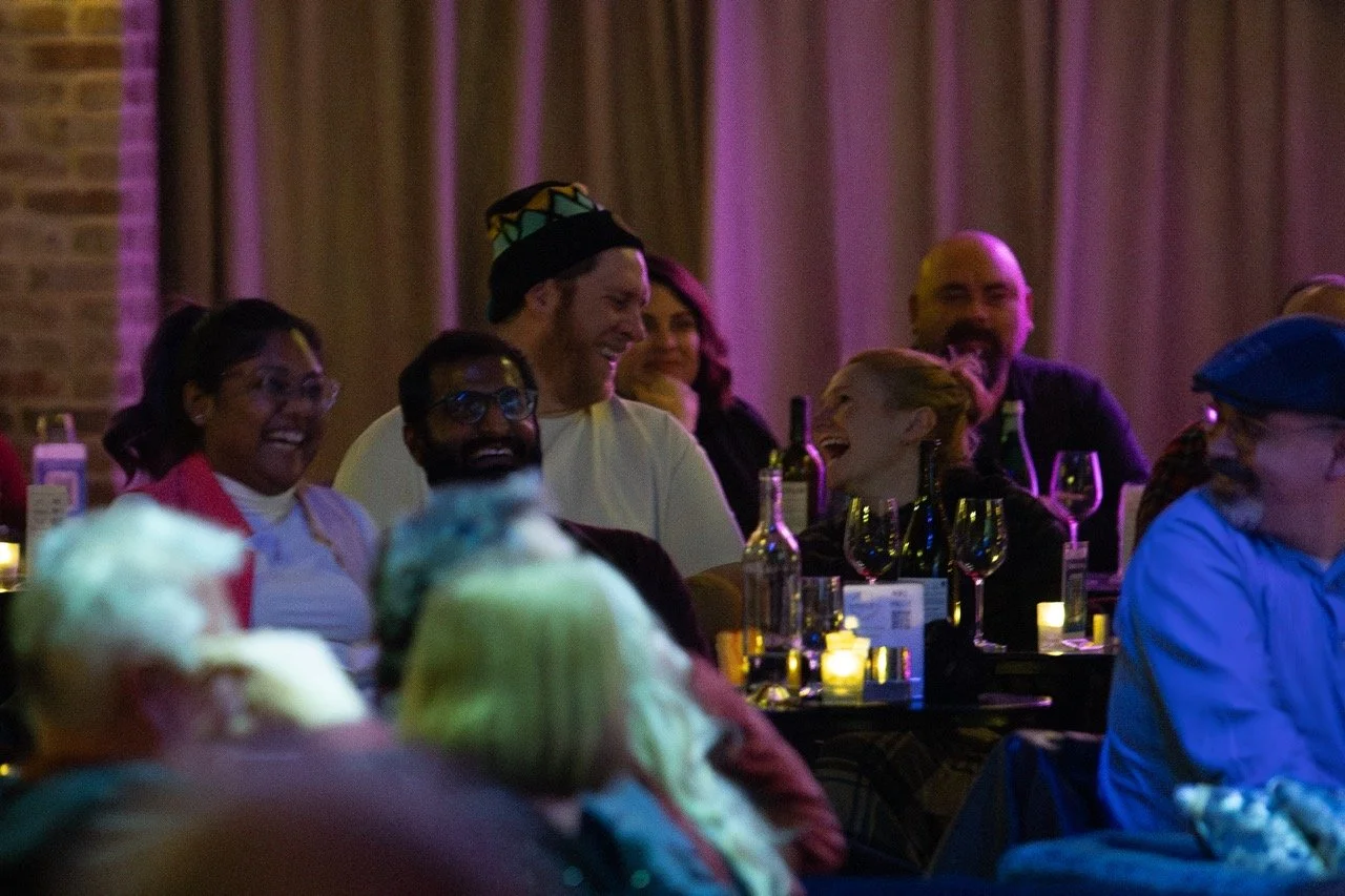 A group of people laughing and enjoying themselves at a social event, with wine glasses and bottles visible on the table, in a dimly lit venue with a warm atmosphere.