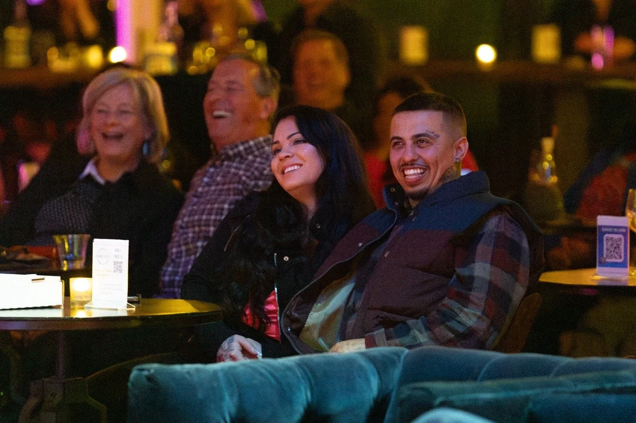 Smiling couple seated with others in a dimly lit indoor venue, enjoying a performance or event.