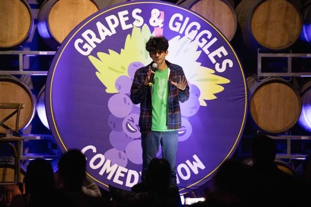 Comedian performing at "Grapes & Giggles Comedy Show," standing on stage with a microphone, barrels in the background.