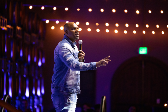 Man speaking on stage with microphone, wearing denim jacket and jeans, under string lights.