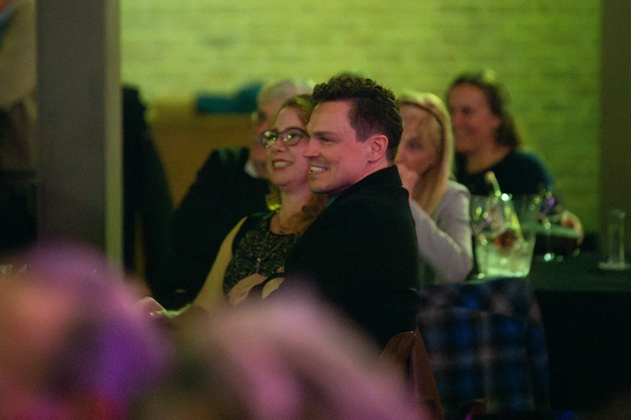 People sitting at a table, smiling and enjoying an event, with a blurred background and a greenish tint.