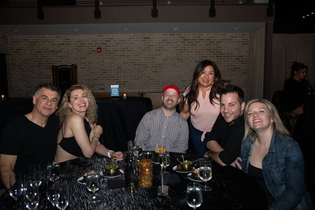 Group of six people sitting and standing around a table with drinks and food in a dimly lit restaurant or venue, smiling at the camera.