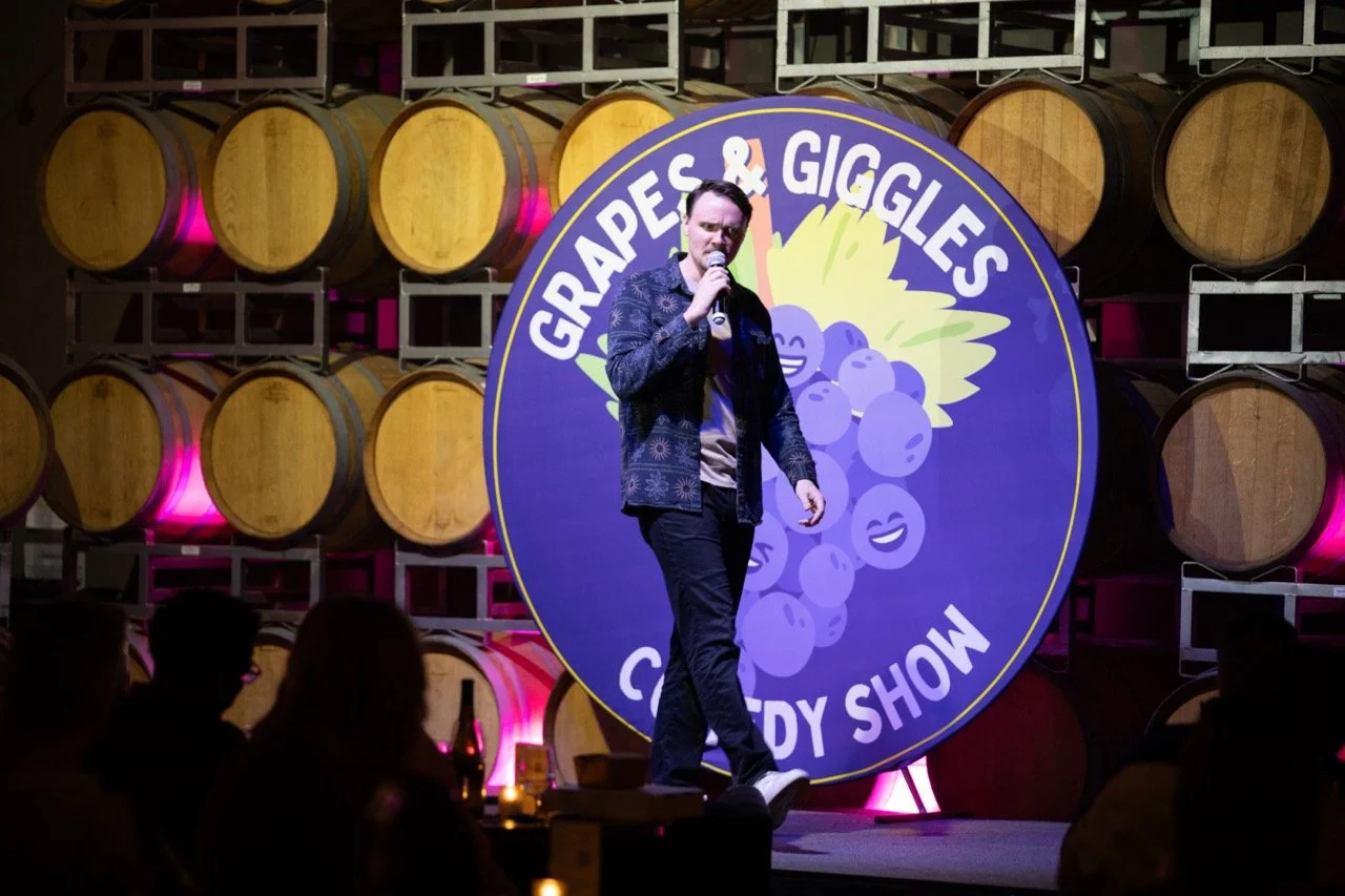 Comedian performing at "Grapes & Giggles Comedy Show" event, with wine barrels in the background.