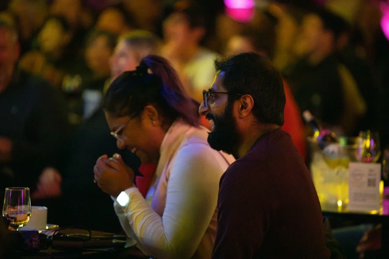 People laughing at an event, sitting at a table with drinks, in a dimly lit room.
