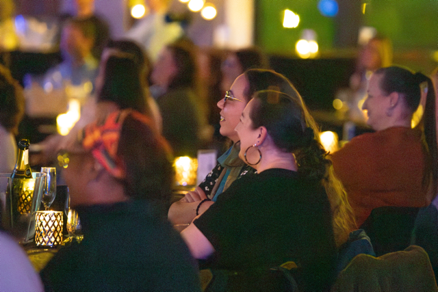 People sitting at tables in a dimly lit venue, likely attending a live event or performance. Candles are on the tables, creating a warm ambiance.