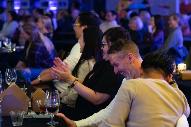 Audience enjoying a comedy show, with people laughing and clapping at tables, wine glasses and food containers visible.