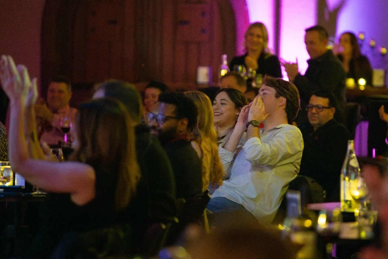 People sitting at tables in a dimly lit venue, watching a performance or event, with a few people clapping and smiling.